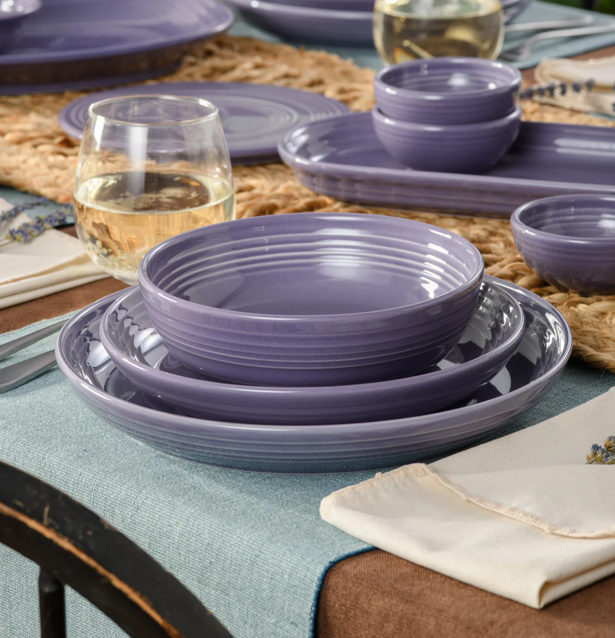 A beautifully set table featuring purple ceramic dishes, a glass of white wine, and linen napkins on a textured table runner.