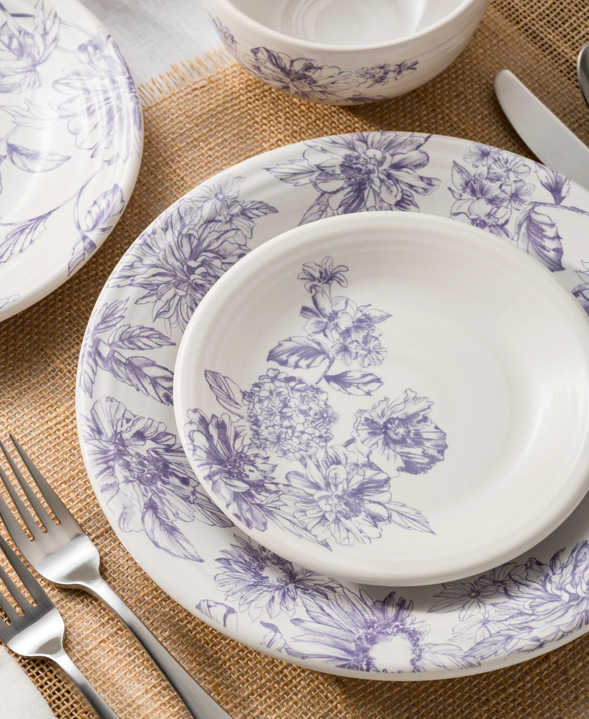 A set of floral-patterned dishes in purple and white, arranged on a burlap table runner with silver cutlery nearby.